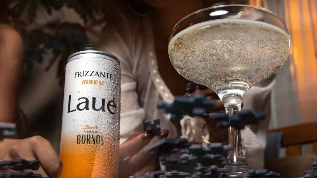 Lata de LAUÉ frizzante fría con gotas de condensación junto a una copa con bebida espumosa, sobre una mesa, mientras una persona juega rompecabezas al fondo en un ambiente íntimo y relajado.