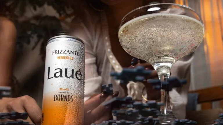 Lata de LAUÉ frizzante fría con gotas de condensación junto a una copa con bebida espumosa, sobre una mesa, mientras una persona juega rompecabezas al fondo en un ambiente íntimo y relajado.