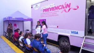 Mujeres sentadas junto a una unidad móvil rosa de Banco Azteca con la identidad de SOMOS Prevención y una carpa de atención.